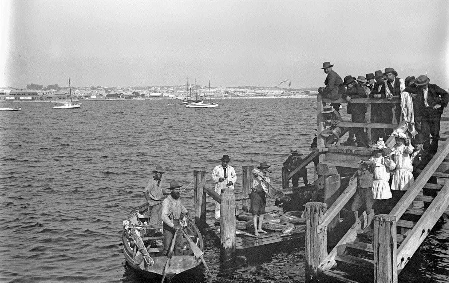 Historic image black and white of fisherman unloading catch onto jetty, moored boats and shoreline in background